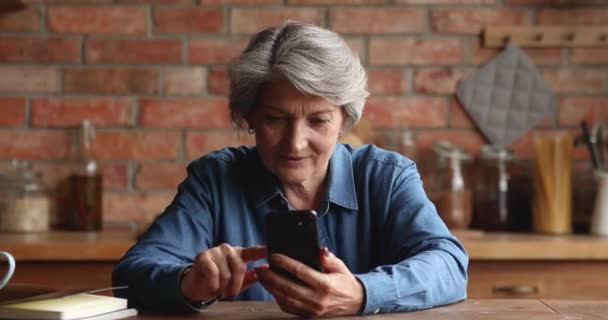 高齢者の女性は、スマートフォンの近代的な無線デバイスとキッチンに座る — ストック動画