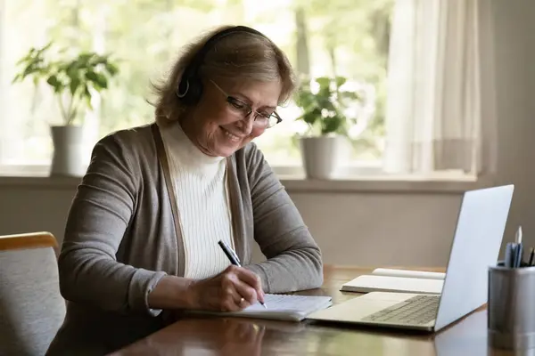 Sorridente donna matura indossando cuffie scrittura note, utilizzando il computer portatile — Foto Stock