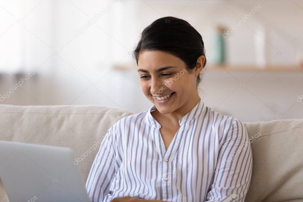 Millennial indian woman sit on couch use laptop install software