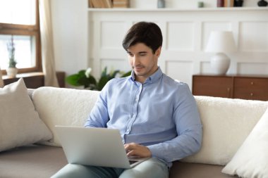 Laptop kullanan odaklanmış bir adam, evdeki koltukta oturuyor.
