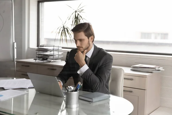 Pensativo hombre de negocios con traje mirando a la pantalla del ordenador portátil, tocando la barbilla —  Fotos de Stock