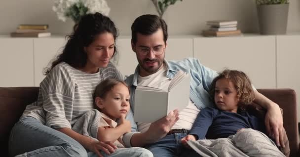 Родители и дети сидят на диване, читая книгу — стоковое видео