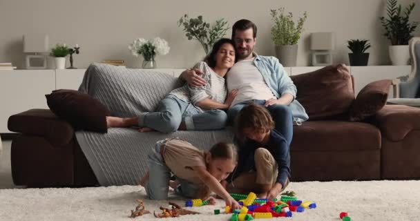 Пара отдыхает на диване, смотрит, как дети играют на полу — стоковое видео