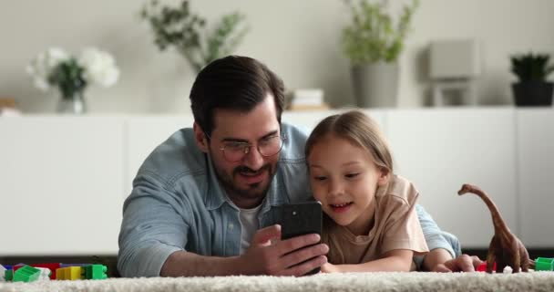 Pai pequena filha deitada no tapete se divertindo usando smartphone — Vídeo de Stock