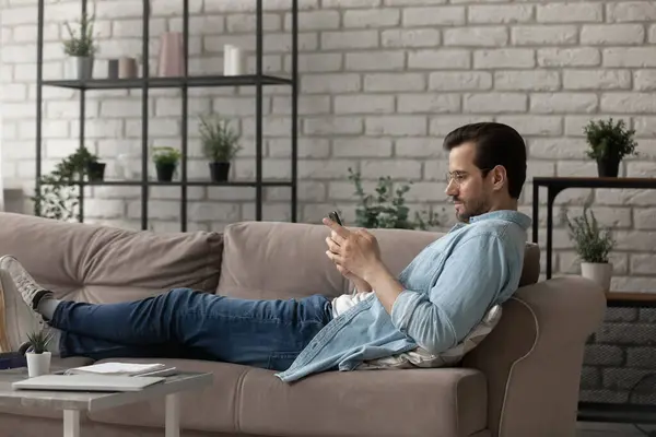 Vista laterale uomo indossa occhiali utilizzando il telefono, sdraiato sul divano — Foto Stock