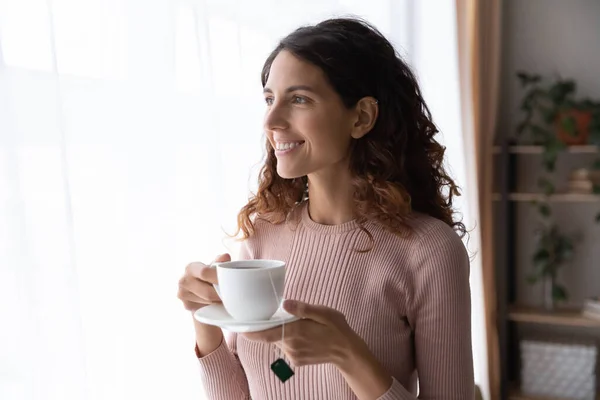 Happy beautiful young dreamy woman standing near window with cup of tea. — Stock Photo, Image