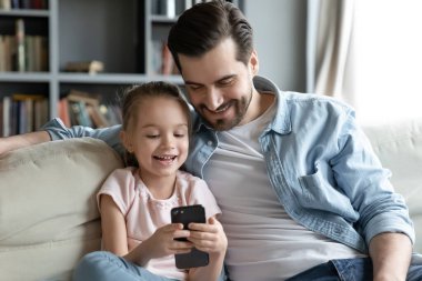 Happy young man cuddling little 6s daughter, playing on smartphone.