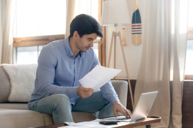 Odaklanmış adam finansal belgeleri kontrol ediyor, dizüstü bilgisayar kullanıyor, bütçeyi planlıyor