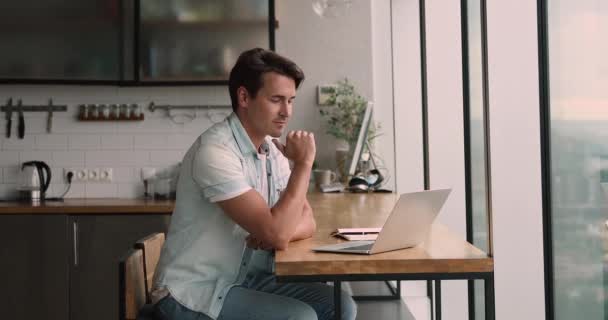 Beschäftigter junger Mann arbeitet am Computer und sitzt in moderner Küche. — Stockvideo