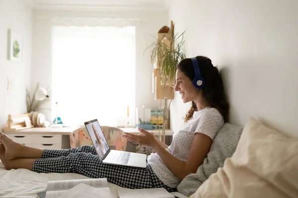 ยิ้มสาวในชุดหูฟังนั่งบนเตียงโดยใช้คอมพิวเตอร์ — ภาพถ่ายสต็อก