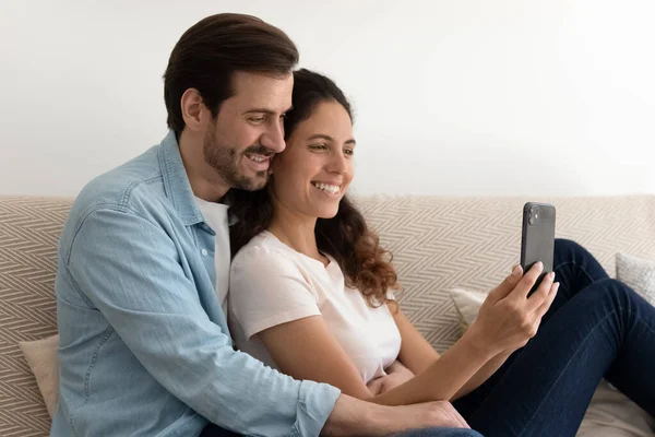 Θετικό παντρεμένο ζευγάρι χαλαρώνει στο σπίτι στον καναπέ κρατώντας κινητό τηλέφωνο — Φωτογραφία Αρχείου