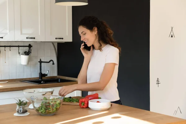 Активная молодая домохозяйка готовит еду на кухне, общаясь по телефону — стоковое фото