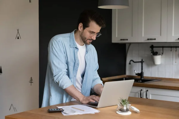 Hombre joven y seguro de proporcionar pagos en línea utilizando el ordenador portátil en la cocina — Foto de Stock