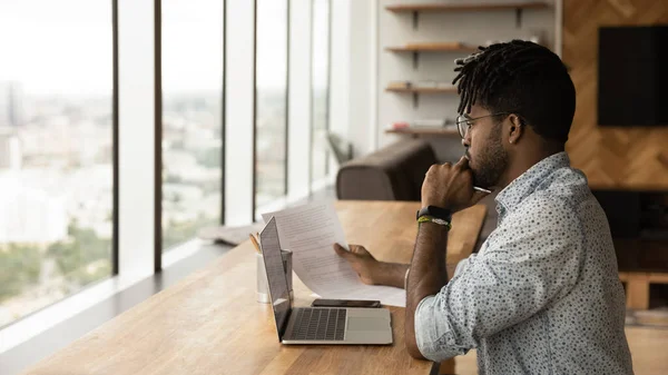 Koncentrerad ung afrikansk man läsa tryckt dokument innan bärbar skärm — Stockfoto