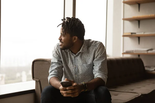 Happy african guy look at window with smartphone in hands — Stock Photo, Image