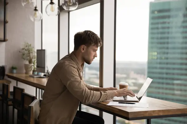 Concentrato uomo millenario utilizzando computer portatile alla scrivania vicino alla finestra panoramica — Foto Stock