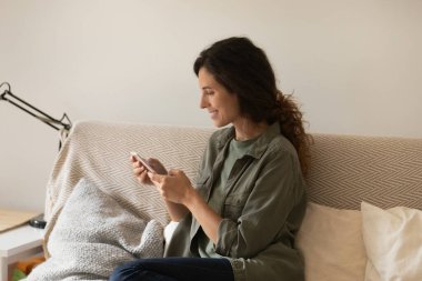 Gülümseyen kadın akıllı telefon kullanarak kanepede rahatlıyor.