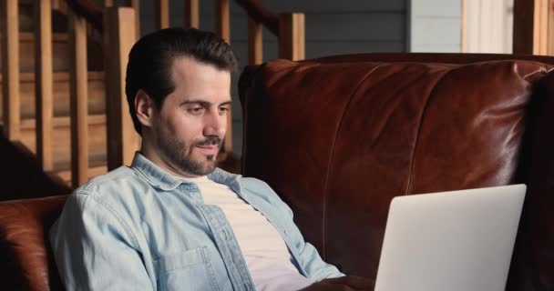 Konzentrierter Millennial-Mann sitzt auf bequemer Couch und arbeitet am Laptop — Stockvideo