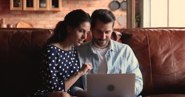 Молодая пара сидит на диване с ноутбуком обсуждая покупку онлайн — стоковое видео