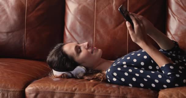 Woman lying indoor listens audio messages on smartphone through headphones — Stock Video