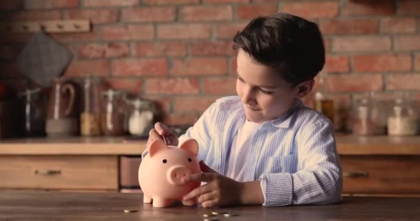 Menino da escola pequeno feliz colocando moedas no banco porquinho. — Vídeo de Stock