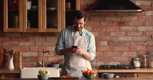 Отвлекает от приготовления пищи улыбающийся молодой человек с помощью смартфона. — стоковое видео