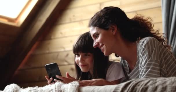 Schattig klein meisje en moeder liggend op bed met smartphone — Stockvideo