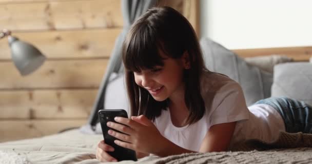 Маленькая девочка лежит на кровати с помощью современного смартфона — стоковое видео