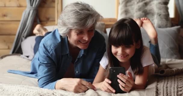 Oudere oma kleine kleindochter liggend op bed met behulp van smartphone — Stockvideo