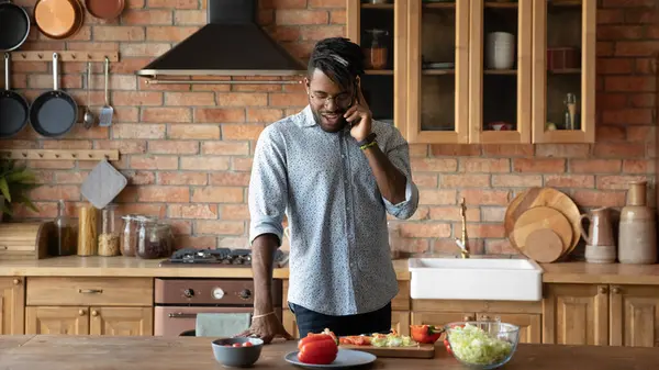 Sonriente hombre afroamericano hablando por teléfono, cocinando en la cocina —  Fotos de Stock