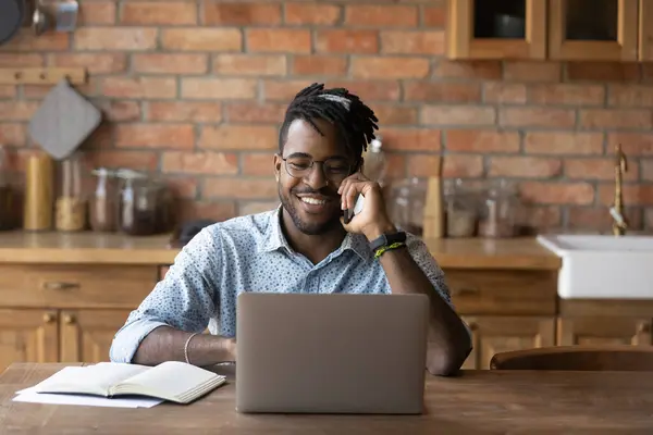 Улыбающийся афроамериканец разговаривает по телефону, используя ноутбук — стоковое фото