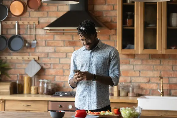 Sourire homme afro-américain en utilisant le téléphone dans la cuisine, la recherche recette — Photo