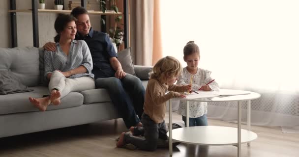 Pareja joven padres descansando, viendo a los niños pequeños dibujar en el álbum. — Vídeo de stock