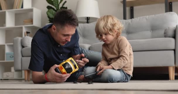 Улыбающийся молодой отец помогает маленькому сыну чинить игрушечную машину. — стоковое видео
