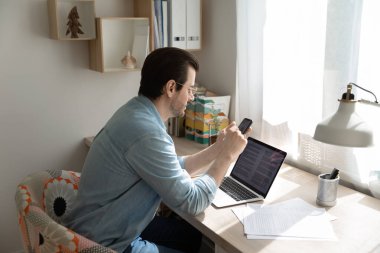 Millennial Man dizüstü bilgisayarda çalışıyor. Akıllı telefondan mesaj atıyor.