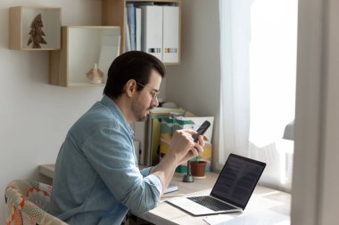 Genç adam akıllı telefon kullanarak evde bilgisayarda çalışıyor.
