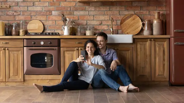 Overjoyed man and woman using phone, sitting on kitchen floor — Stock Photo, Image