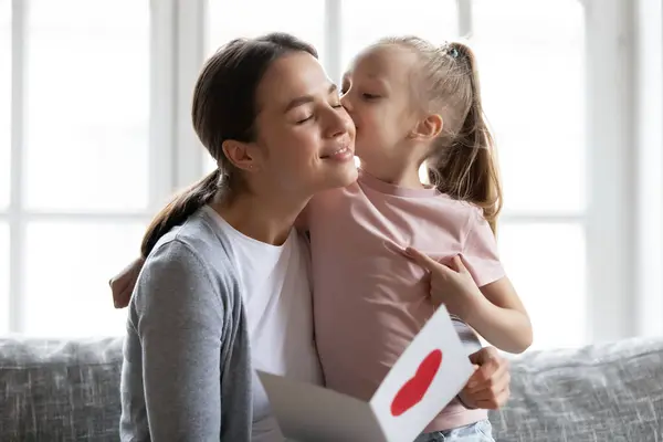 Linda hija niño besando a mamá y dando tarjeta de felicitación hecha a mano —  Fotos de Stock