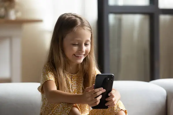 Happy small girl sit on sofa play game using cellphone — Stock Photo, Image