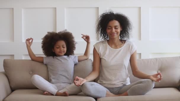 African mom teaching funny kid daughter meditate together on sofa — стокове відео