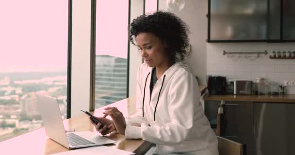 Distraído del trabajo en la computadora joven mujer africana feliz usando teléfono inteligente. — Vídeos de Stock