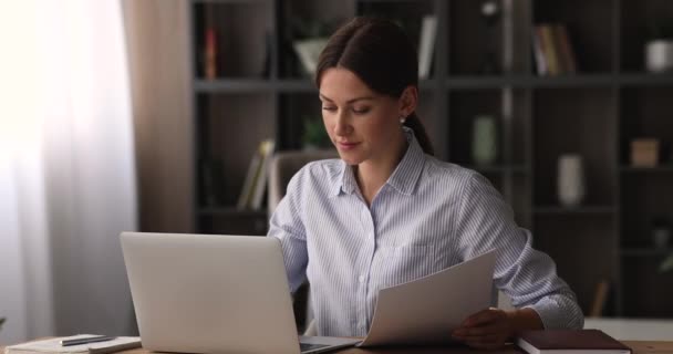 Εστιασμένη νεαρή επιχειρηματίας που εργάζεται σε φορητό υπολογιστή, αναλύοντας έγγραφα σε χαρτί. — Αρχείο Βίντεο