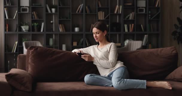 Joyeux jeune femme utilisant des applications de téléphone portable, reposant sur le canapé. — Video