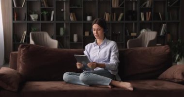 Kulaklıklı rahat genç bir kadın elinde tablet kullanarak video çağrısı tutuyor..