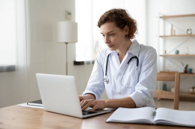 Genç beyaz kadın doktor internette dizüstü bilgisayarda çalışıyor.