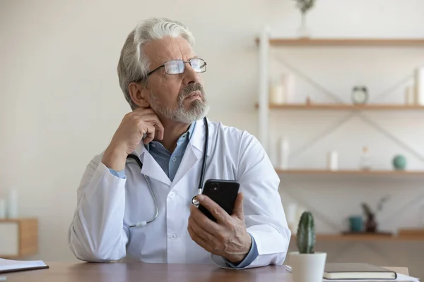 Pensive senior male doctor use mobile thinking — Stock fotografie