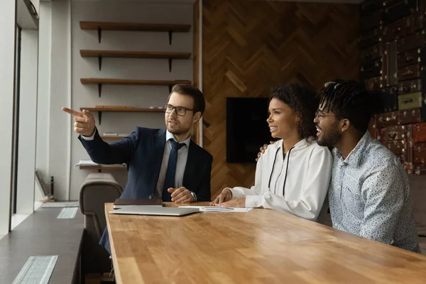 Inmobiliaria caucásica consultando a pareja afroamericana, apuntando a la ventana — Foto de Stock