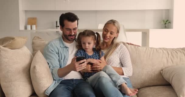 Пара и дочь отдыхают на диване, наслаждаясь смартфоном — стоковое видео
