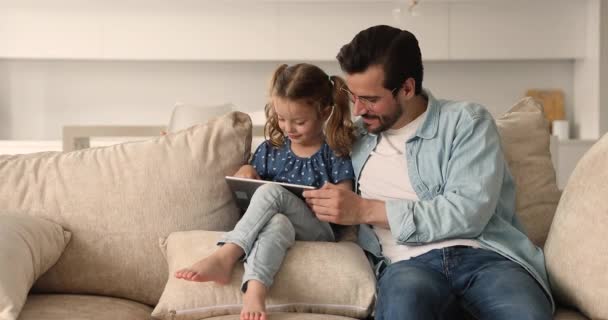 Fader sin lilla dotter sitta på bekväm soffa med hjälp av surfplatta — Stockvideo