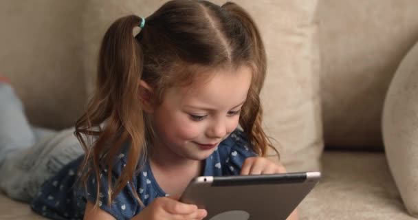 Klein meisje liggend op bank holding tablet spelen online games — Stockvideo